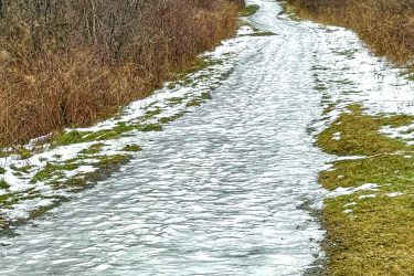 A narrow winding path covered in a thin layer of ice and patches of snow, flanked by bare trees and brush. The scene is set against a cloudy sky, hinting at a chilly, winter day. The path leads further into the distance, suggesting a tranquil outdoor setting. Iroquois Heights mountain bike trail.