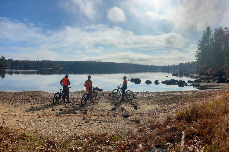 Three mountain bikers, wearing bright orange vests, stand by a tranquil lake on a sunny day, facing the water. The reflections of trees and clouds can be seen on the lake's surface, and the shoreline is lined with rocks and autumn foliage.