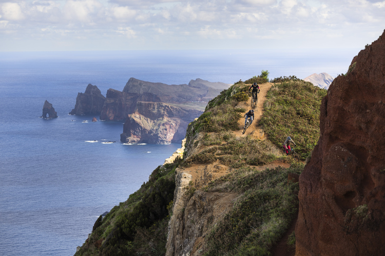 Three mountain bikers ride along a narrow trail on a hillside overlooking the ocean, with rugged cliffs and rock formations in the background. The sky is partly cloudy, and the scene captures a mix of lush greenery and sandy earth, highlighting the dramatic coastal landscape.