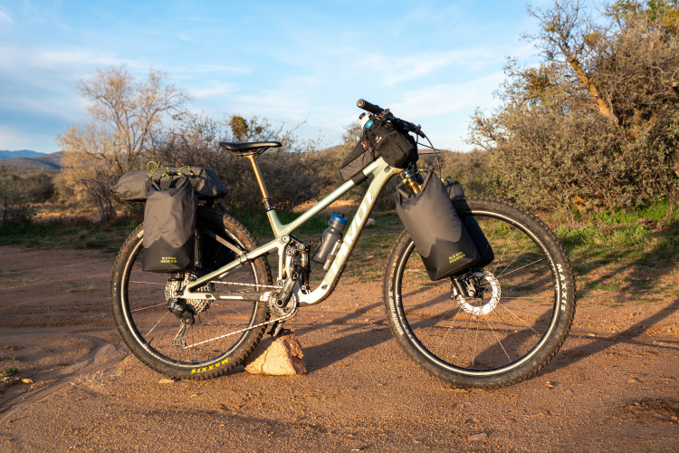 A mountain bike equipped with black travel bags is parked on a dirt path surrounded by sparse vegetation and distant hills under a clear blue sky. The bike features a green frame and is oriented towards the right, showcasing its components, including a water bottle holder and backpack-style attachments.