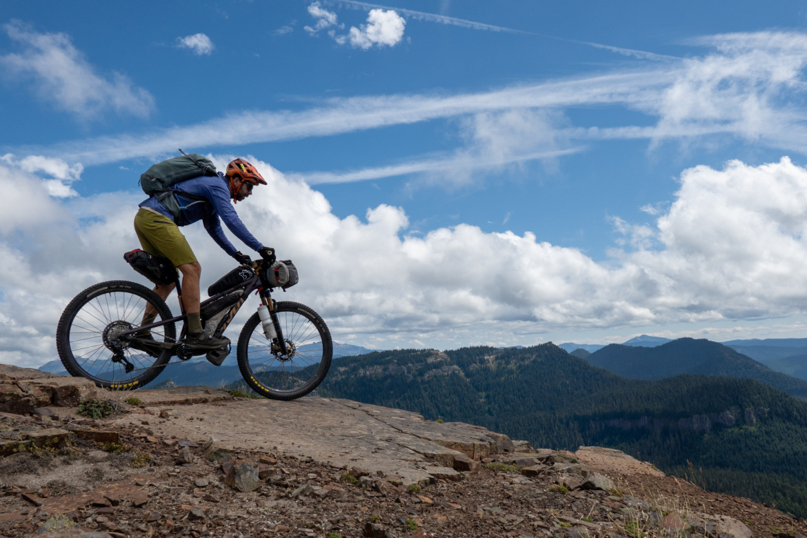 A cyclist navigates a rocky trail at a high elevation, with mountains and a blue sky dotted with clouds in the background. The rider is wearing a helmet and carrying a backpack, focusing intently on the path ahead.