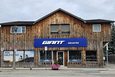 A wooden storefront with the sign "GIANT ANCASTER" prominently displayed in blue. Large windows feature bicycles on display, and a nearby lamppost and flowering plants add to the scene. The sky is overcast, suggesting a cloudy day.
