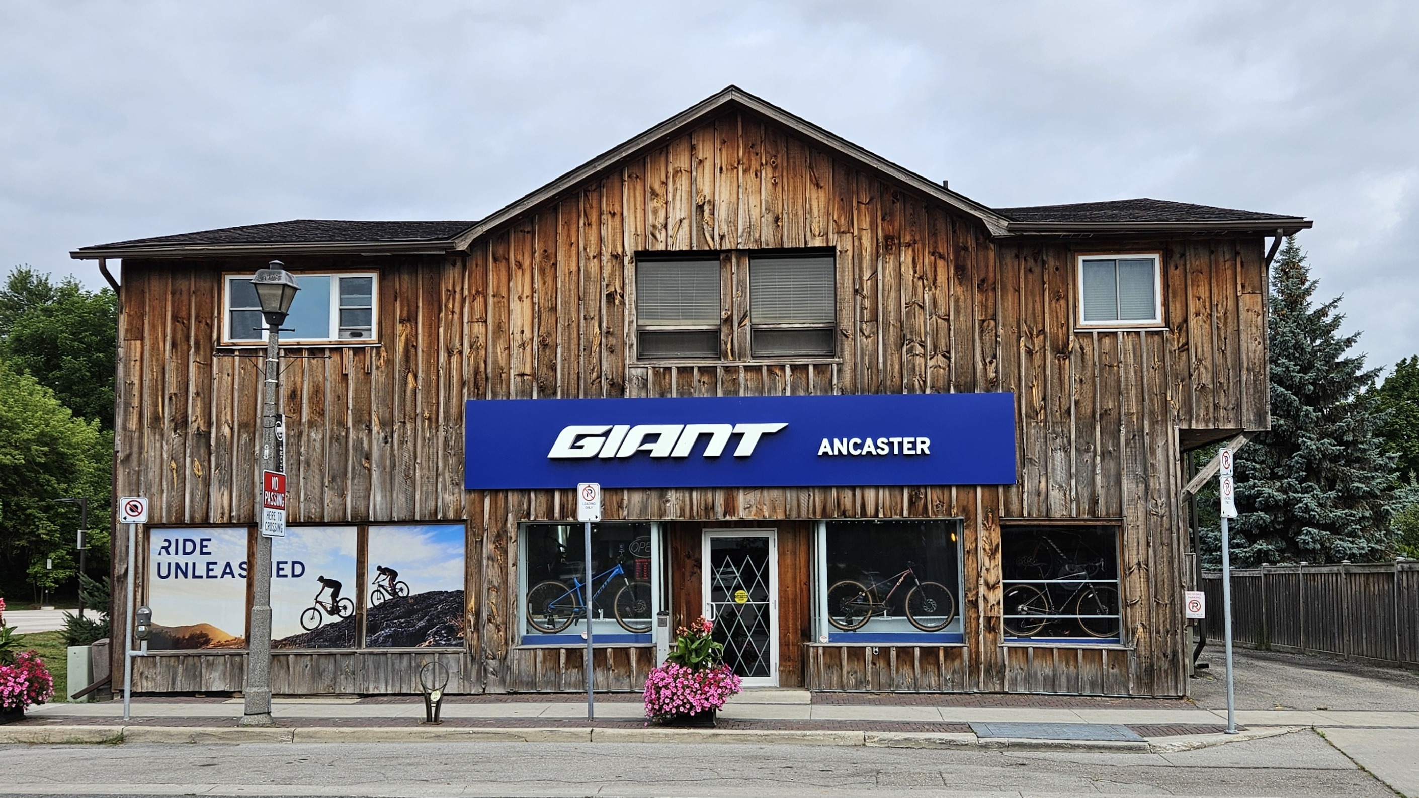 A wooden storefront with the sign "GIANT ANCASTER" prominently displayed in blue. Large windows feature bicycles on display, and a nearby lamppost and flowering plants add to the scene. The sky is overcast, suggesting a cloudy day.