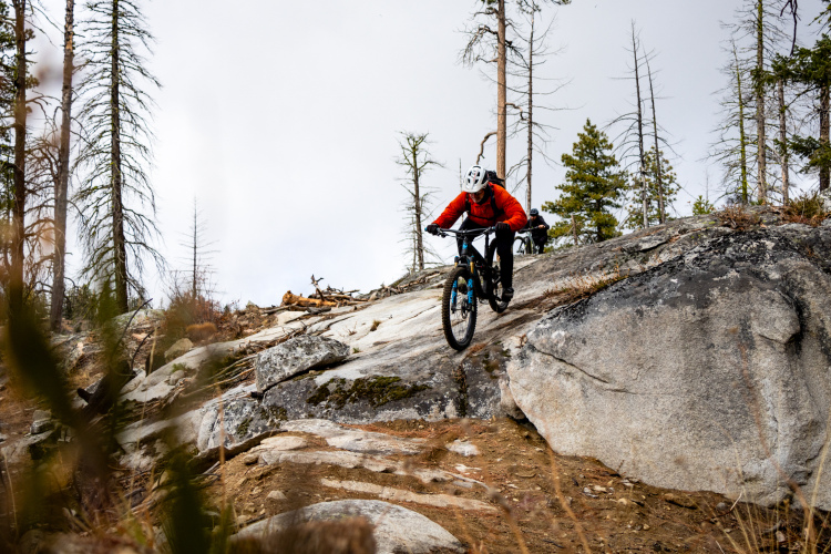 A mountain biker in a red jacket descends a rocky slope, navigating rugged terrain surrounded by pine trees. Closer to the camera, another cyclist can be seen riding in the background, highlighting the adventurous outdoor setting.