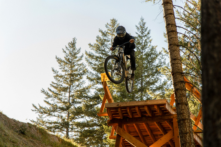A person in a black riding outfit and helmet performs a jump on a mountain bike off a wooden ramp in a forested area. Tall evergreen trees surround the scene, and the sky is clear in the background.