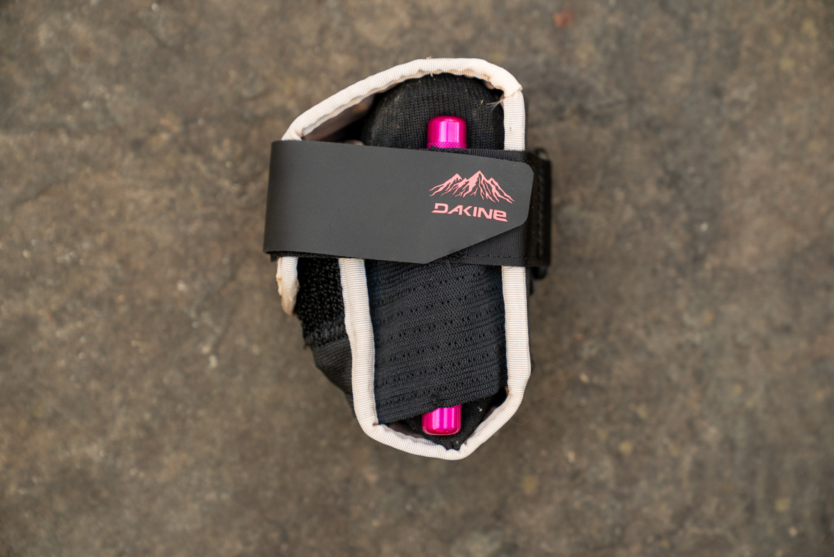 A close-up view of a black and beige Dakine wrist guard or protective gear, featuring a pink logo and a purple accessory peeking out from inside. The item is displayed on a textured surface.