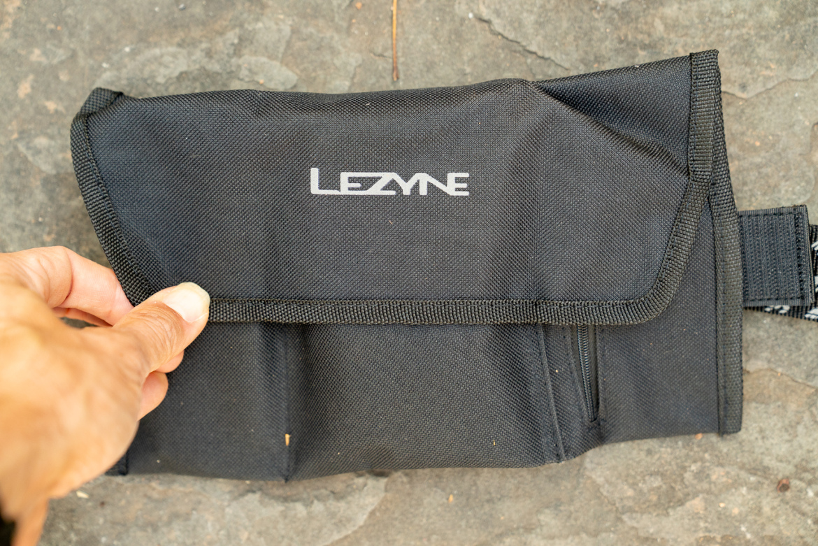 Black fabric pouch with a flap, featuring the "Lezyne" logo on the front. A hand is seen holding the pouch, which is placed on a textured surface.