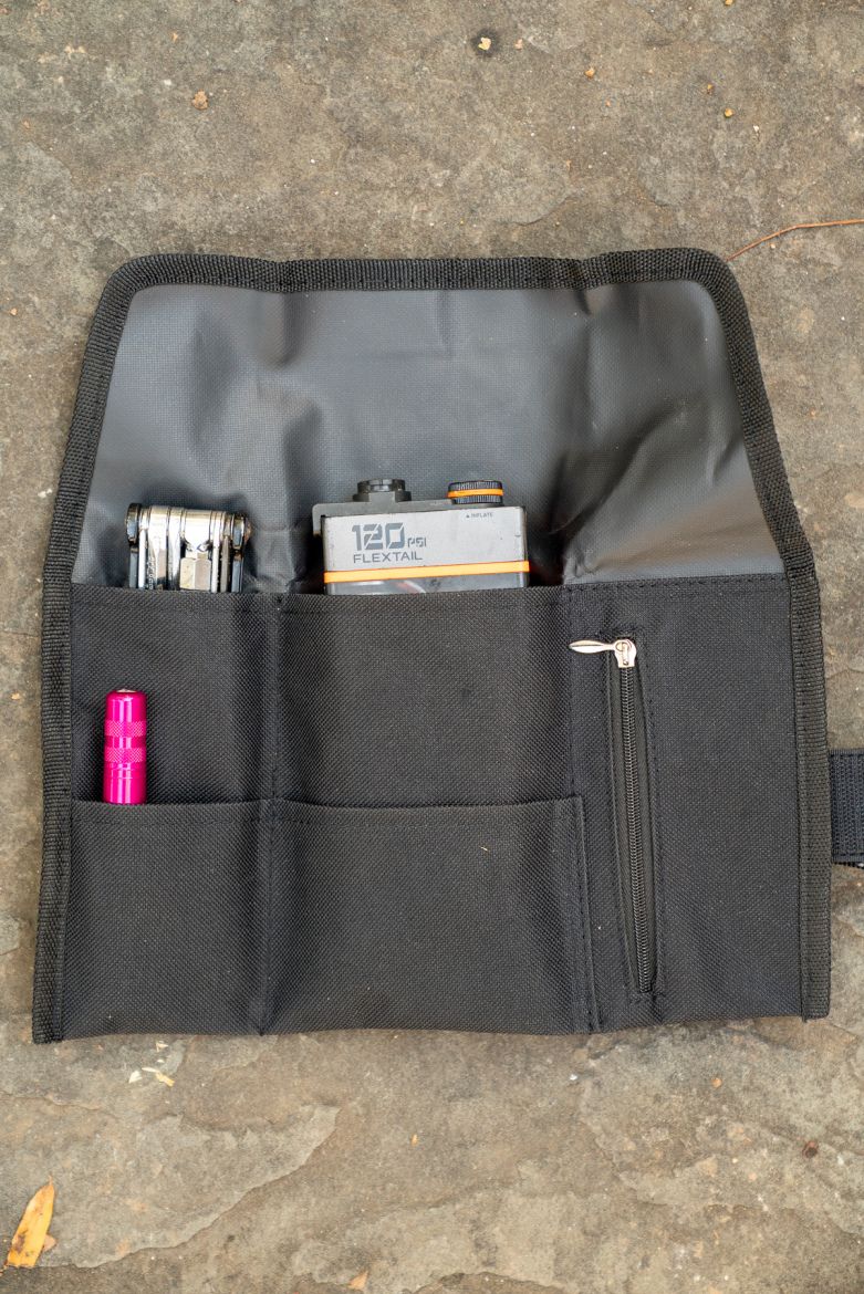 A black organizer pouch laid flat on a rocky surface, containing a pink flashlight, a multi-tool, and a device labeled "120 PSI FLEXTAIL." The pouch features multiple compartments to hold various tools and accessories.