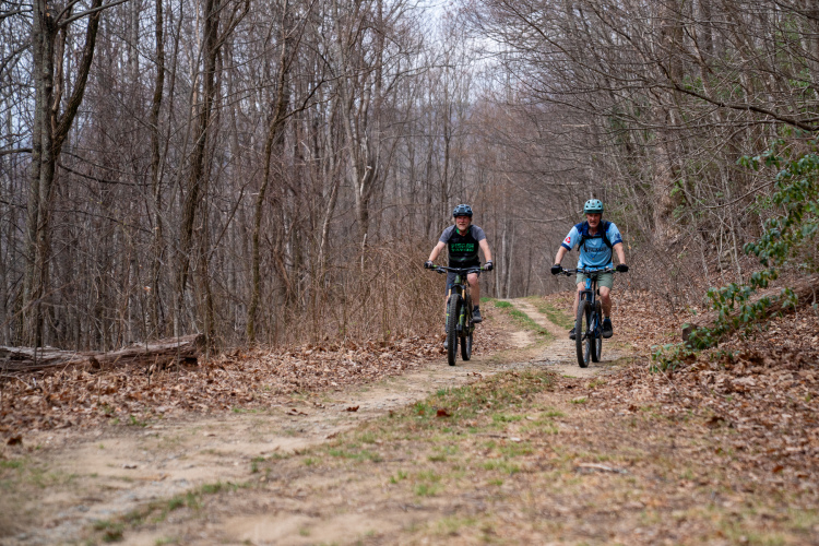 Two mountain bikers riding along a dirt trail in a wooded area during early spring. The trees are bare, and fallen leaves are scattered along the path. The bikers, wearing helmets and casual athletic clothing, appear to be enjoying their ride on a cool, overcast day.
