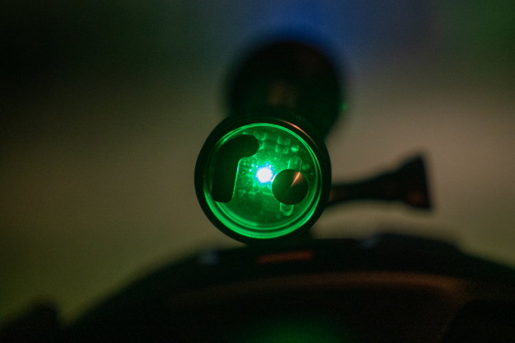 A close-up view of a green flashlight lens illuminated by a blue light, set against a blurred, dark background. The lens features a circular design with a small blue light in the center, and a metal body is visible behind it.