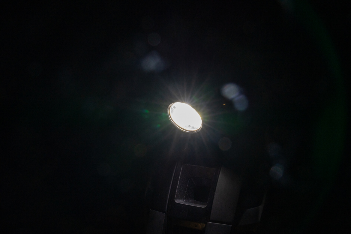 A close-up view of a bright light source shining in the darkness, creating rays and reflections. The light is circular and is mounted on a dark surface, with hints of lens flare effects visible around it.