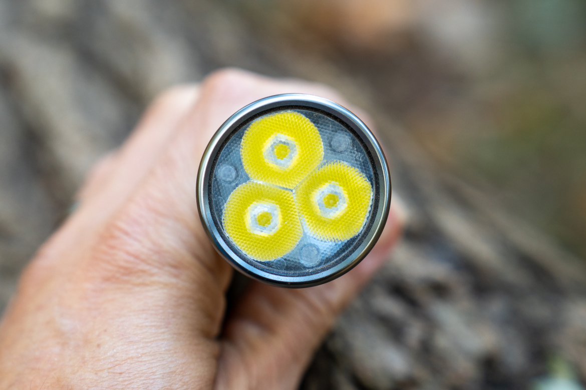 Close-up of a person's hand holding a flashlight, showcasing the light's lens with three yellow LEDs arranged in a triangular formation. The background is blurred, revealing a natural outdoor setting with hints of brown and green.