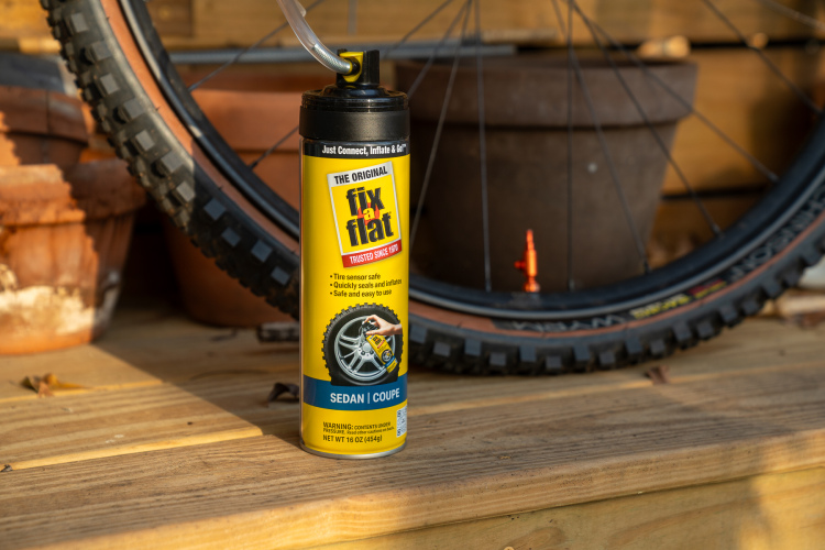 A canister of Fix-a-Flat tire repair product is positioned on a wooden surface, with a bicycle wheel partially visible in the background. The can features a bright yellow design with text highlighting its use for sedans and coupes, and directions for easy inflation and sealing. Potted plants are also seen in the background, adding to the outdoor setting.