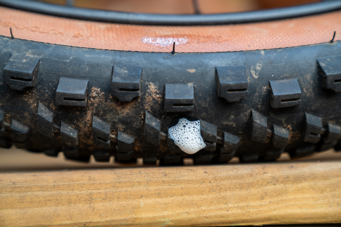 Close-up of a mountain bike tire resting on a wooden surface, showing dirt and a small area of foam near the tread, indicating potential sealant leakage.