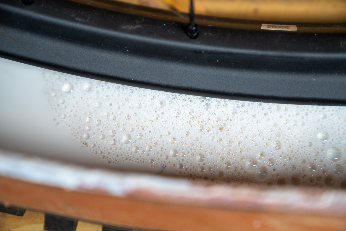 Close-up view of soap suds in a round basin, with a black rim at the top edge and wooden surface visible below. The bubbles vary in size, creating a frothy texture.