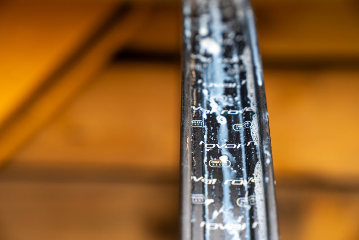 A close-up view of a bike rim with water and soap bubbles, featuring branding and markings that indicate it is tubeless ready. The background is softly blurred, highlighting the rim's surface texture.