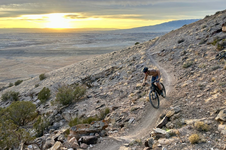 A mountain biker navigates a winding trail on a hillside, surrounded by rocky terrain and sparse vegetation. The sun sets in the background, casting a warm glow over the expansive landscape below.
