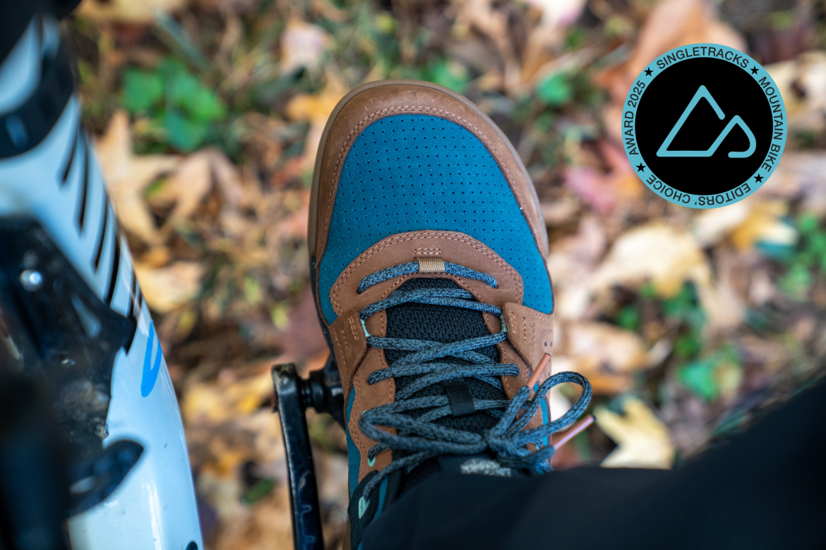 Close-up of a cycling shoe featuring a brown and blue design, positioned on a bike pedal against a background of autumn leaves. The image includes a circular logo in the corner indicating an award for mountain bike gear.