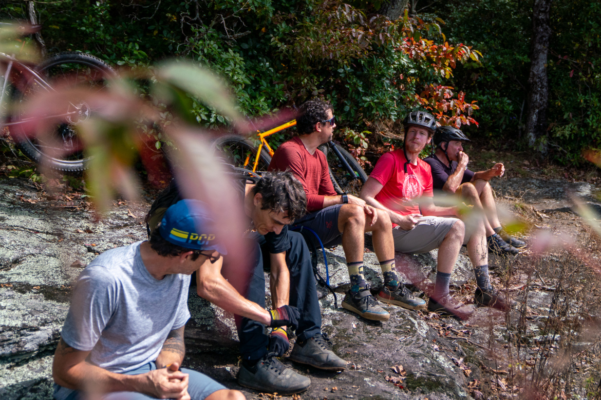 A group of five mountain bikers sitting on a rocky surface among lush greenery. They are taking a break, chatting, and adjusting their gear. Two bikes are visible in the background. The scene captures a sunny day with colorful foliage around them.