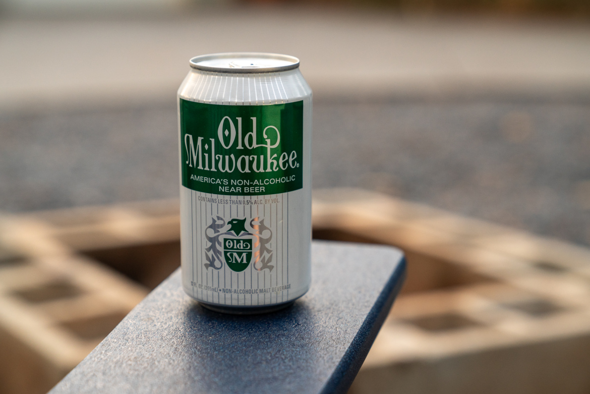 A can of Old Milwaukee non-alcoholic beer sitting on a blue table in an outdoor setting. The can features green and white branding with the text "Old Milwaukee" and "America's Non-Alcoholic Near Beer." The background includes a blurred view of a stone surface.