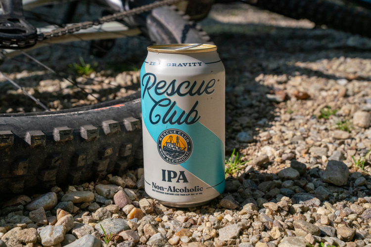 A can of Rescue Club Non-Alcoholic IPA is positioned on a gravel surface, with a bicycle tire partially visible in the background. The can features a blue and silver design, prominently displaying the brand name and product type.