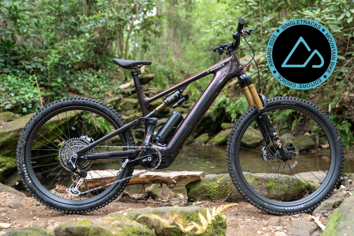 A high-performance mountain bike is positioned on a rocky path near a stream, surrounded by lush greenery. The bike features a sleek, dark frame with high-quality components, including a prominent shock absorber and wide, rugged tires suitable for off-road terrain. In the corner of the image, there is a logo indicating a mountain bike award for 2025 from Singletracks.