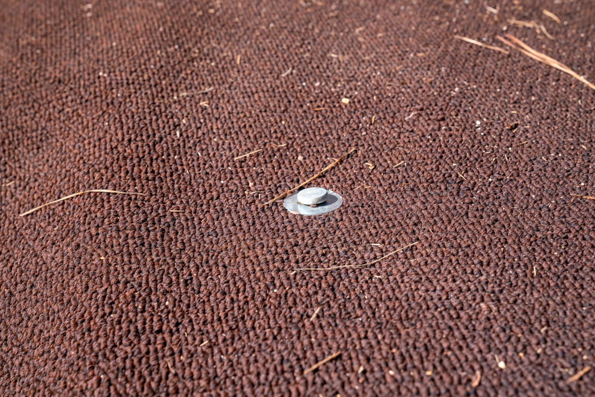 A close-up view of a textured brown fabric surface with a metal fastener positioned in the center. Small twigs and debris are scattered across the fabric.