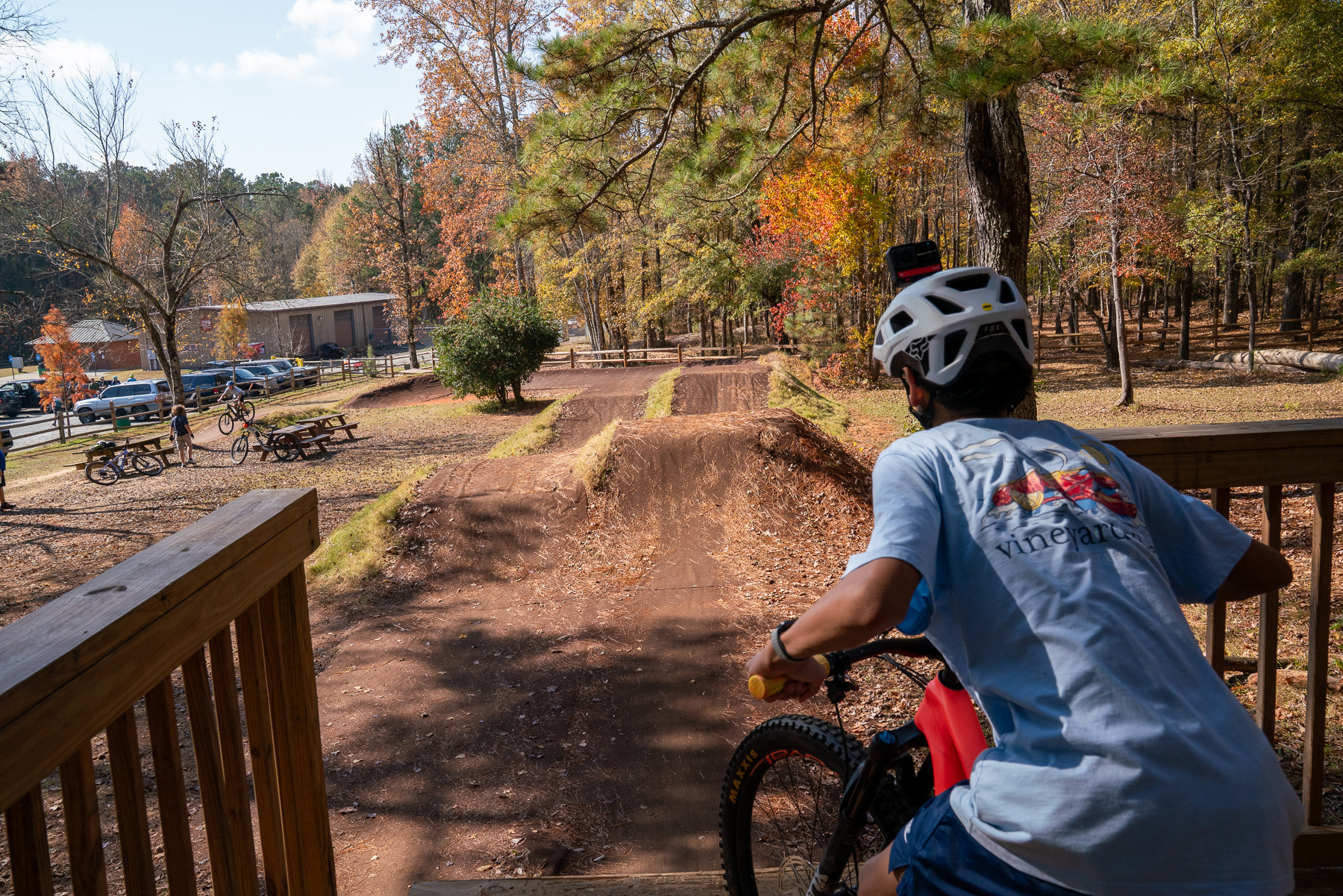 A person in a helmet sits on a mountain bike at a wooden deck, looking down a dirt biking path with jumps and ramps, surrounded by autumn foliage. In the background, there are picnic tables and other bikers enjoying the area.