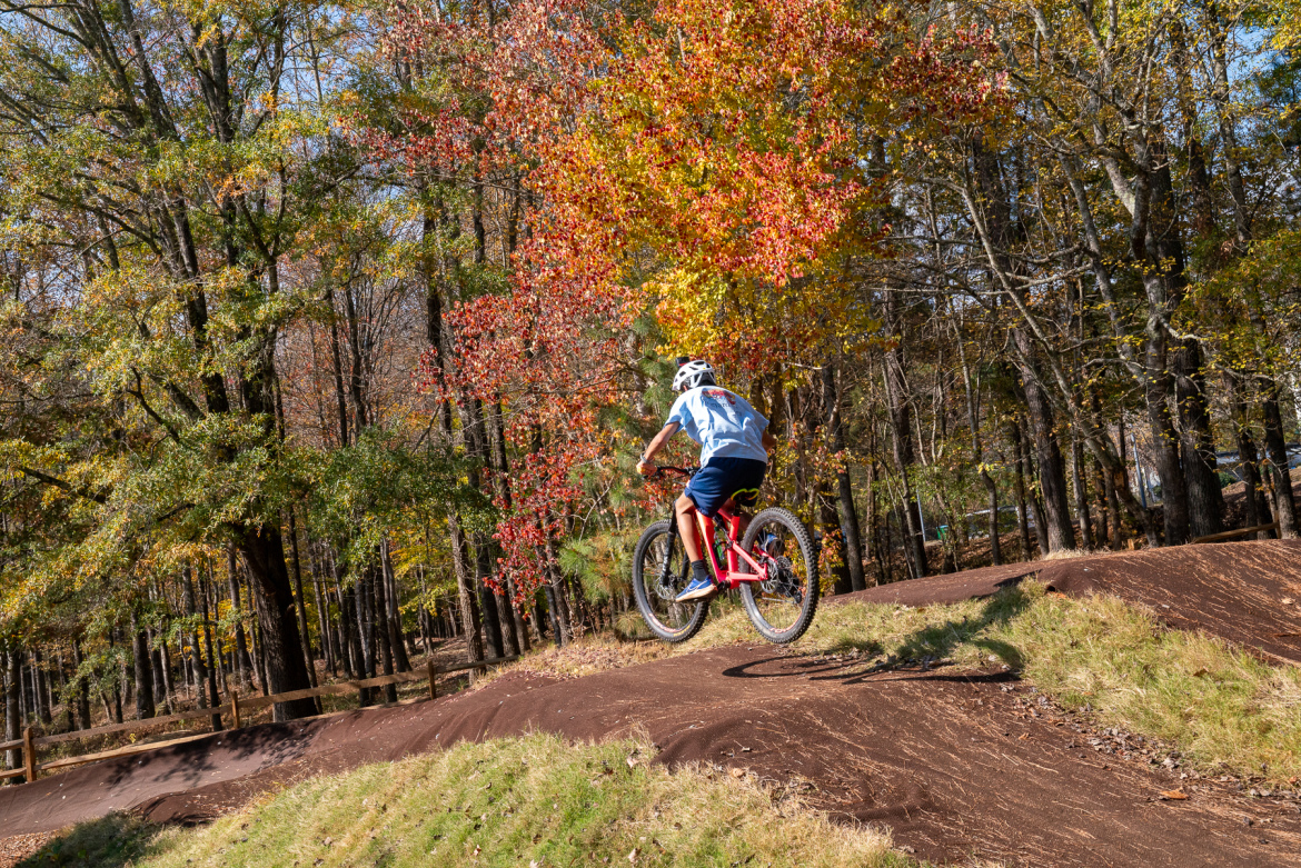 A person in a blue shirt and shorts rides a pink mountain bike, jumping over a dirt ramp in a forested area with autumn foliage in vibrant shades of red and orange. The scene captures a moment of action in an outdoor biking environment.