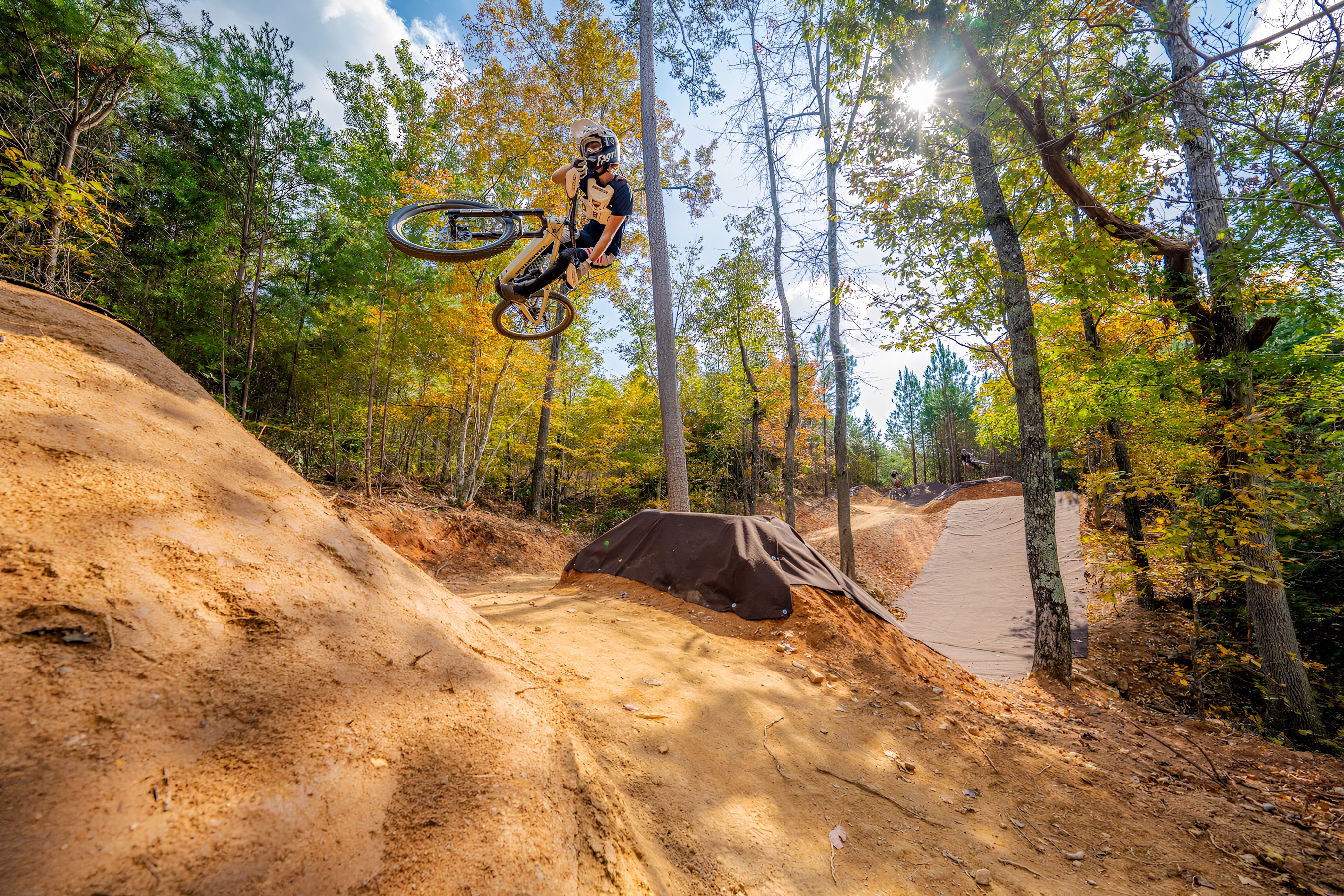 A cyclist in mid-air performing a trick on a dirt bike jump surrounded by trees with autumn foliage. The sun is shining through the branches, casting light on the dirt bike trail in the background. Sendy Crawford mountain bike trail.