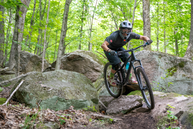 A mountain biker skillfully navigating rocky terrain in a lush, green forest. The rider is wearing a helmet and protective gear, leans forward on the bike, and maneuvers over large rocks and dirt paths surrounded by trees.
