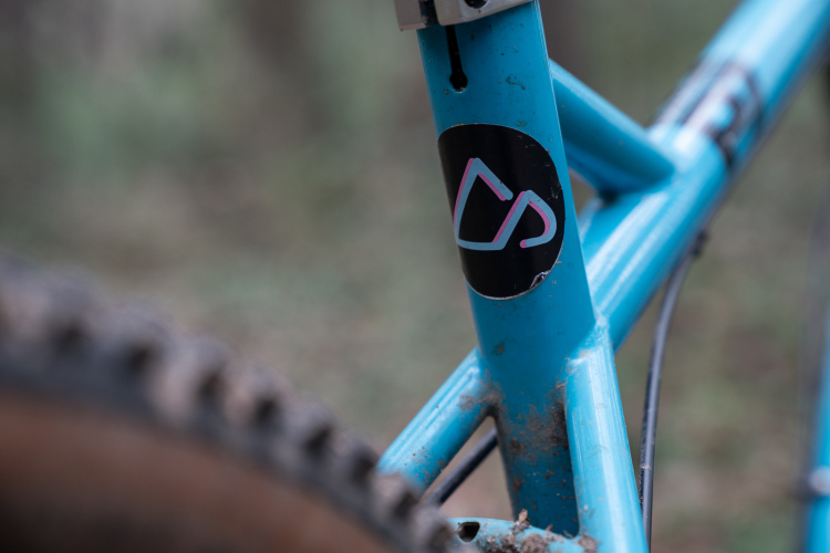 Close-up of a blue bicycle frame with a black sticker featuring a stylized logo, set against a blurred natural background. The frame shows signs of outdoor use with some dirt visible.