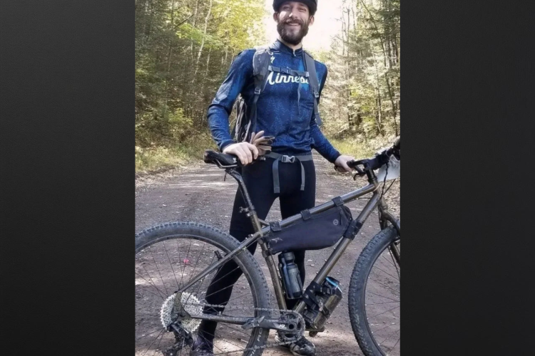 A man stands on a dirt trail beside his mountain bike, wearing a helmet and a long-sleeved blue shirt with "Minnesota" printed on it. He has a backpack on, black leggings, and is smiling at the camera. The background shows a forested area with trees and sunlight filtering through the leaves.