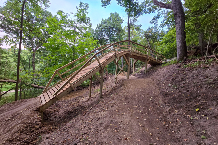A wooden bridge arches over a sloping path in a lush green forest. The bridge is supported by several wooden beams and leads up a dirt trail, surrounded by trees and underbrush. The sky is slightly cloudy, and the scene is filled with natural light.