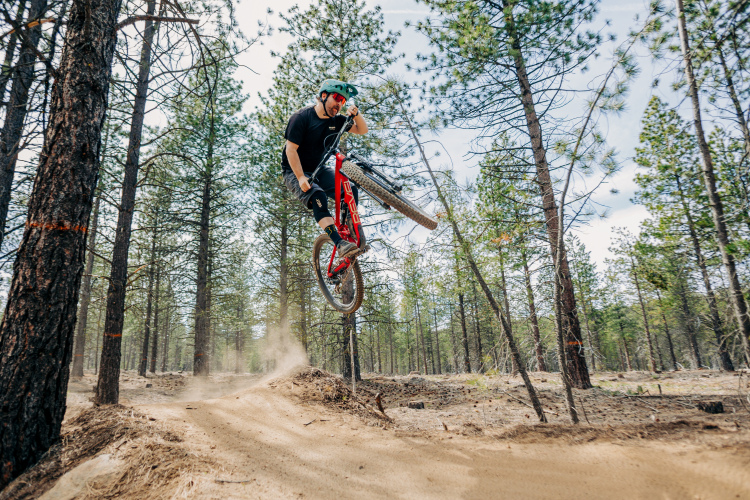 A mountain biker performing a jump on a dirt trail surrounded by tall pine trees, kicking up dust as he catches air above the ground. The rider is wearing a helmet and protective gear, showcasing a dynamic pose mid-air.