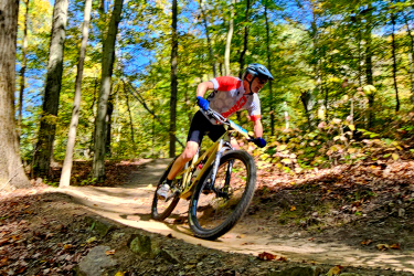 A mountain biker navigates a dirt trail through a colorful autumn forest, leaning into a turn with determination. The scene features tall trees with vibrant green and yellow leaves under a clear blue sky. The rider is dressed in a red and gray jersey, blue gloves, and a helmet. West Branch mountain bike trail.