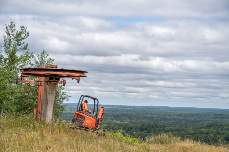 Construction worker operating a small excavator near an old ski lift structure, with a panoramic view of green forests and cloudy skies in the background.