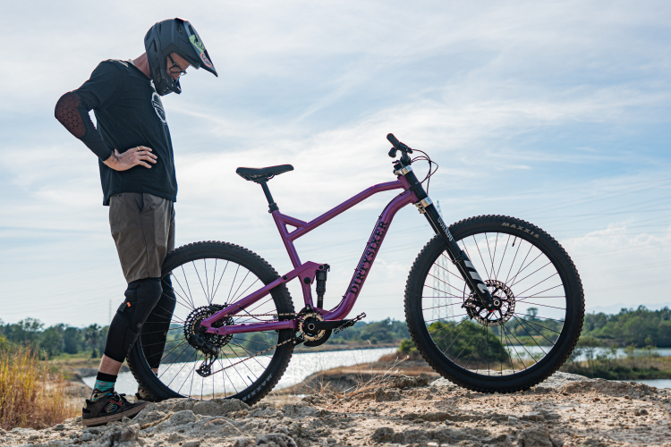 A mountain biker in protective gear stands beside a purple mountain bike on rocky terrain, gazing down at the bike. The background features a scenic view of water and trees under a partly cloudy sky.