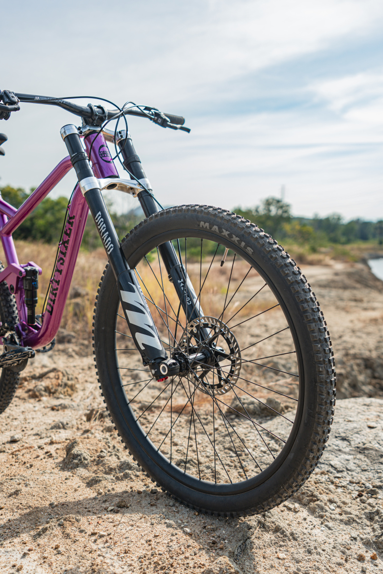 Close-up of a mountain bike's front wheel and fork, showcasing a purple frame, black suspension fork, and tire with "Maxxis" branding. The bike is positioned on sandy terrain with a blurred background of greenery and a body of water under a clear sky.