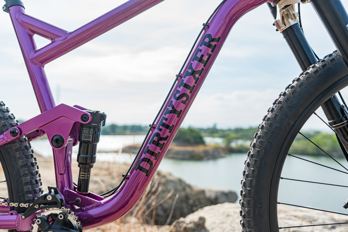 Close-up view of a purple mountain bike frame featuring the name "DIRTYSIXER" prominently displayed. The image highlights the bike's suspension system, including the RockShox shock, as well as the rugged tire and surrounding natural landscape near a body of water.