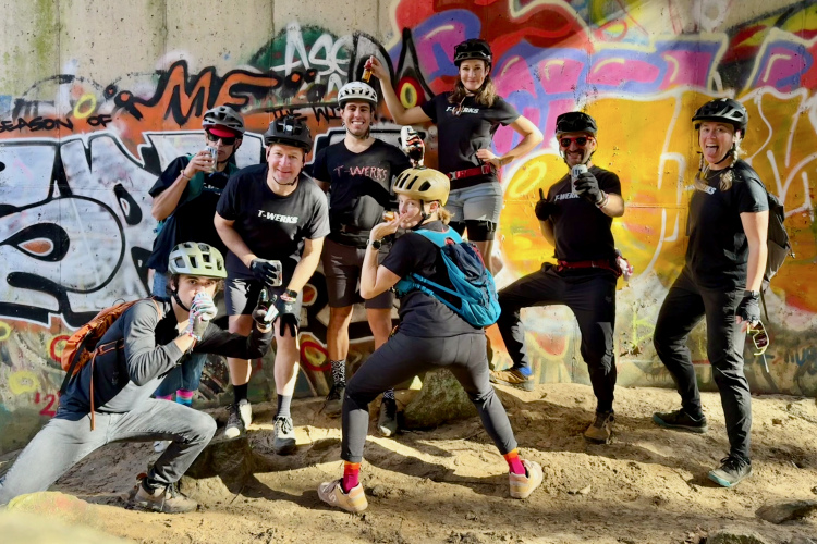 A group of eight people posing joyfully in front of a colorful graffiti wall, wearing cycling helmets and casual athletic attire. They are in a sandy area, with some participants crouching or leaning in playful stances, while others hold drinks and smile at the camera.