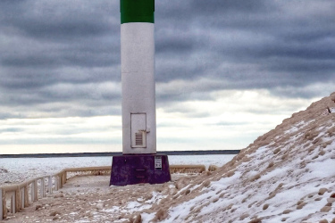 A lighthouse standing on a snowy shore, with a green top and a white base. The path leading to the lighthouse is covered in snow and ice, under a cloudy sky. The Beach mountain bike trail.