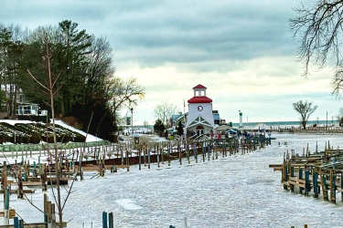 A winter scene showing a partially frozen marina with empty docks lined on either side. In the distance, a building with a red roof and a clock tower is visible, surrounded by trees and a cloudy sky. The icy water stretches toward the horizon, with a few boats visible in the distance. The Beach mountain bike trail.