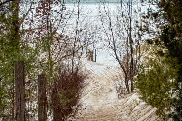 A winding dirt path leads through a sparse, wintry landscape, bordered by bare trees and shrubs. In the background, a calm body of water is visible beneath a cloudy sky, creating a serene atmosphere. The path, partially covered in snow, suggests a tranquil outdoor scene inviting exploration. Pinery Provincial Park mountain bike trail.