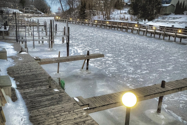 A winter scene depicting a frozen canal with wooden docks on either side. The foreground features a wooden walkway leading along the water's edge, while a lamp post with a glowing orb illuminates the area. Snow covers the ground, and bare trees line the canal, under a cloudy sky. The Beach mountain bike trail.
