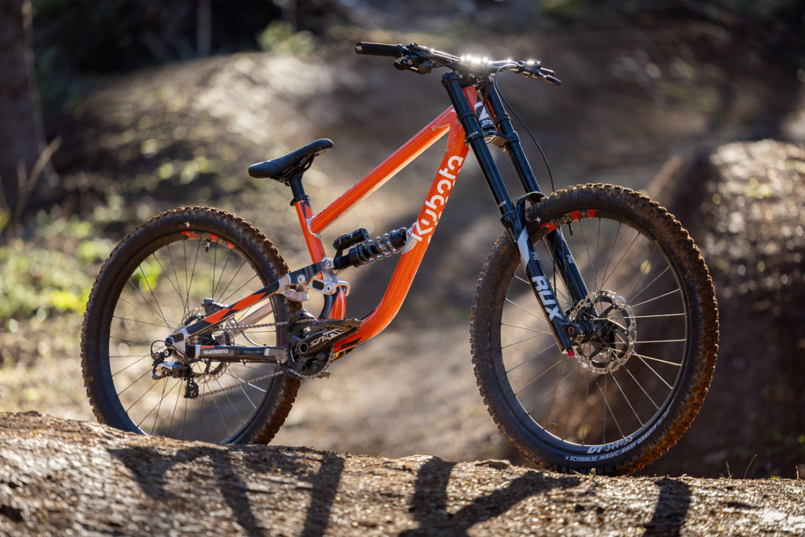 A vibrant orange and black mountain bike positioned on a dirt trail, with the front wheel resting on a log and a blurred forest background. The bike features a sturdy frame and thick tires, suitable for off-road riding.
