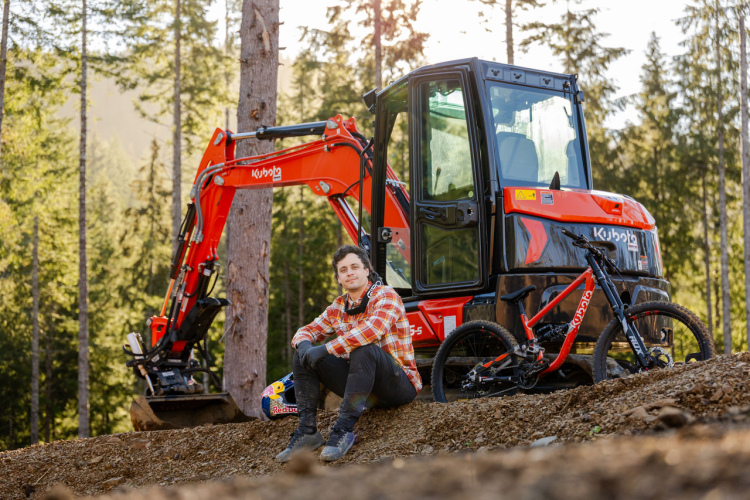 A person in a plaid shirt and gloves sits on a dirt pile next to a Kubota excavator in a forested area. A mountain bike is leaning against the dirt nearby, and trees are visible in the background, suggesting an outdoor adventure or construction scene.