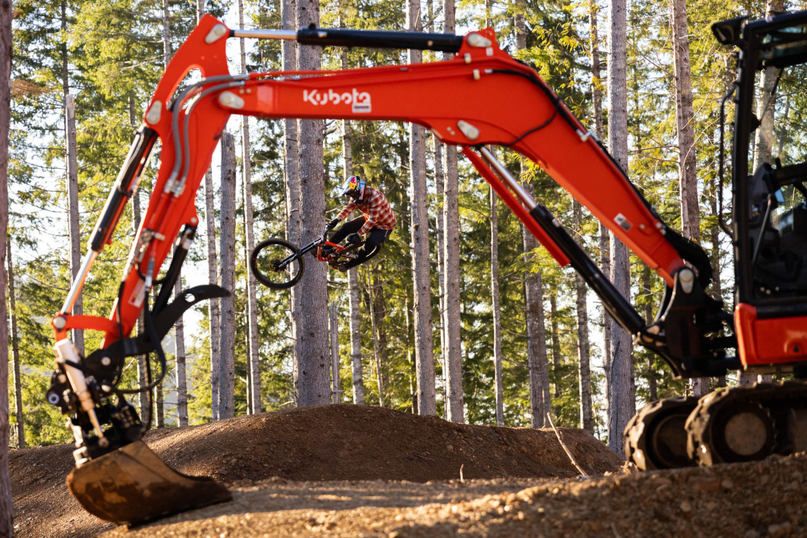 A mountain biker performing a jump over a dirt mound, with a bright orange excavator arm in the foreground. Tall trees surround the scene, highlighting the outdoor setting. The biker is wearing a helmet and a red plaid shirt, showcasing their dynamic movement in mid-air.