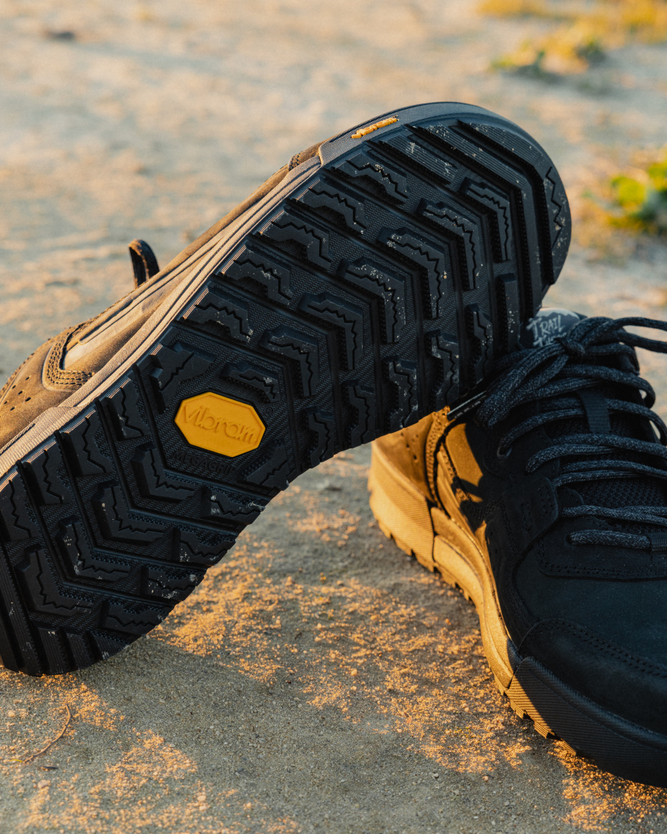 A close-up image of a pair of athletic shoes on a sandy surface, highlighting the textured black outsole with a distinct Vibram logo. The shoes feature a combination of black and tan colors, with laces prominently displayed.