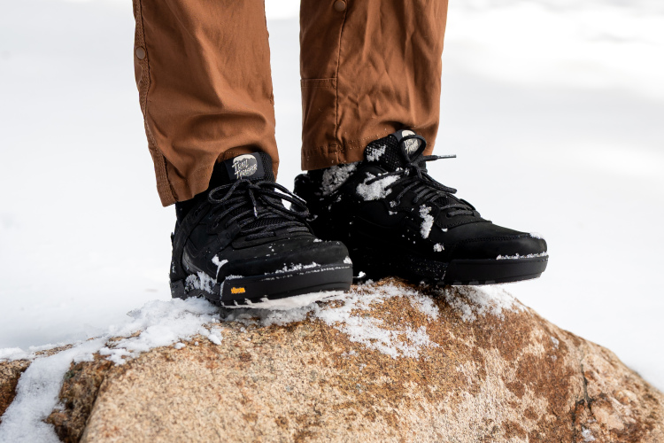 A close-up of black sneakers, partially covered in snow, positioned on a large rock. The person wearing the shoes is dressed in brown outdoor pants, with a snowy landscape in the background.